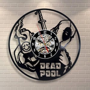 May include: Black vinyl record clock featuring a Deadpool design. The clock has a black face with white numbers and a visible mechanical movement. The word "DEADPOOL" is cut out at the bottom of the clock.