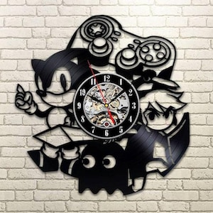 Peut inclure: Horloge murale noire sur le thème des jeux vidéo. Le design intègre des personnages emblématiques et une silhouette de manette. Le cadran est noir avec des chiffres blancs et des mécanismes internes visibles. Les aiguilles sont rouges.