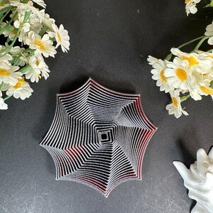 Op de afbeelding: Een geometrisch, stervormig decoratief object met zwarte en zilveren strepen en rode accenten. Het object heeft een vierkant midden en is omgeven door witte en gele bloemen op een zwarte achtergrond.