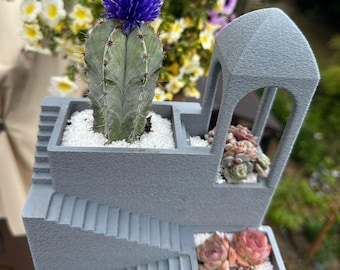 May include: A gray concrete planter with a staircase design, holding a cactus with a blue flower and succulents. The planter has multiple levels and is filled with white pebbles. The background features colorful flowers.