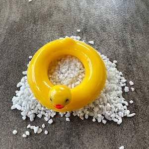 Peut inclure: Un anneau de bouée de piscine jaune vif en forme de canard, avec un bec rouge et des yeux noirs, posé sur un lit de petits cailloux blancs. Un design joyeux pour l'été.