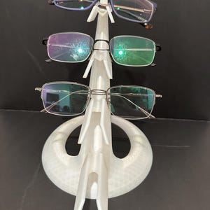Könnte beinhalten: Ein weißer, 3D-gedruckter Brillenhalter mit drei Brillen. Der Halter hat ein einzigartiges Design mit einem kreisförmigen Sockel und einer vertikalen Struktur. Die Brillen haben unterschiedliche Rahmenstile und -farben.