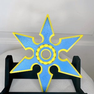 May include: A blue and yellow six-pointed star-shaped fidget spinner with a ring of yellow beads in the centre. The spinner is on a black stand.