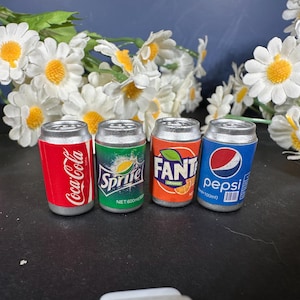 May include: Miniature soda cans, including Coca-Cola, Sprite, Fanta Orange, and Pepsi, are displayed. The cans are silver with colorful labels. The background features white flowers with yellow centers and green leaves.
