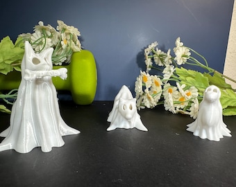 May include: Three white 3D-printed figurines of animals in ghost costumes. The figurines are a fox, a cat holding a bone, and an owl. The figurines are on a black surface with white flowers and a green dumbbell in the background.