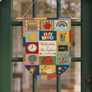 May include: A rectangular fabric wall hanging featuring a patchwork design with colorful illustrations of school supplies, a rainbow, a globe, and a clock. The center panel displays the text Welcome to Ms. Sophia's CLASSROOM with two red hearts.