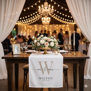 May include: A wedding reception table with a white runner bearing "Mr & Mrs" and the name "Wilton". A floral centrepiece, candles, and champagne flutes are arranged on the dark wooden table. A framed photograph is also present.