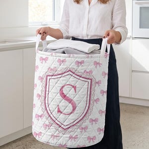 May include: A white quilted laundry hamper featuring a repeating pattern of small pink bows and a central crest design containing a large pink letter S. The cylindrical hamper has two white fabric handles and contains folded white fabric items.