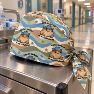 May include: A scrub cap featuring a repeating pattern of brown toads wearing blue cowboy hats. The text "hold on partner" and "im overstimulated" is printed near each toad. The background consists of wavy blue, green, and tan stripes with small stars.