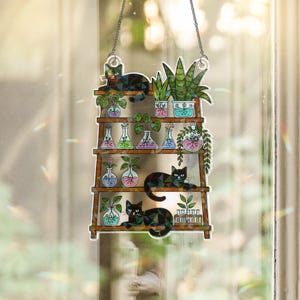 May include: A decorative window hanging featuring a wooden shelf design with plants and black cats. The shelf holds various potted plants, including succulents and hanging vines, and several black cats are lounging on the shelves. The artwork is suspended by a chain.