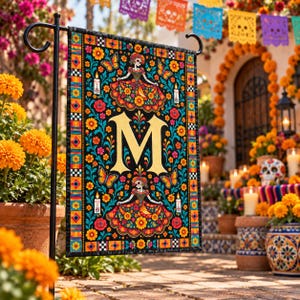 May include: A rectangular garden flag featuring a black background with a large gold letter M in the centre. The design includes two skeletal figures in colourful dresses, orange marigold flowers, butterflies, and white candles, all framed by a vibrant border.