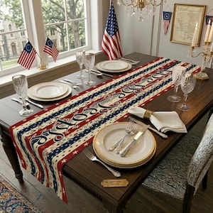 May include: A patriotic table setting with a red, white, and blue Independence Day themed table runner. The runner features stars and stripes with the word "Liberty" in a script font. Includes plates, silverware, and glassware.
