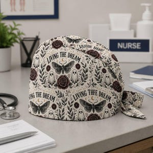 May include: A scrub cap with a cream background featuring a repeating pattern of dark moths, ghosts, and roses. The text "LIVING THE DREAM" is printed in black lettering throughout the design. The cap includes a tie-back closure on the side.