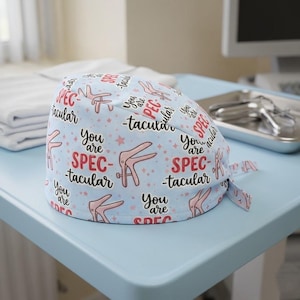 May include: A light blue medical scrub cap with a repeating pattern of pink medical instruments, stars, and the words "You are Spectacular" in red and black. The cap is tied at the back.