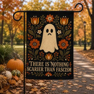 May include: A black garden flag featuring a central cream-colored ghost illustration surrounded by orange and brown floral patterns. The text "THERE IS NOTHING SCARIER THAN FASCISM" is printed in a bold, serif font at the bottom of the rectangular flag.