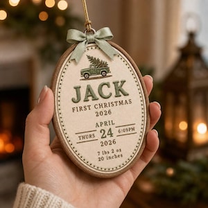 Könnte beinhalten: Ein ovales, besticktes Ornament in einem Holzrahmen mit einem grünen LKW, der einen Baum trägt, dem Namen JACK und Geburtsdaten: 24. April 2026, 18:09 Uhr, 3,23 kg und 50,8 cm, verziert mit einer salbeigrünen Schleife.