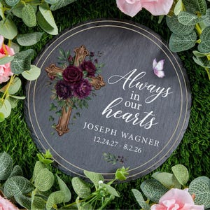 May include: A round, dark gray memorial stone with a gold-colored cross adorned with burgundy flowers. The text "Always in our hearts" and a name and dates are printed in white. A butterfly and floral accents complete the design.