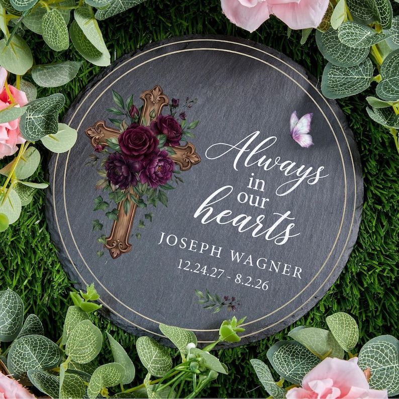 May include: A round, dark gray memorial stone with a gold-colored cross adorned with burgundy flowers. The text "Always in our hearts" and a name and dates are printed in white. A butterfly and floral accents complete the design.