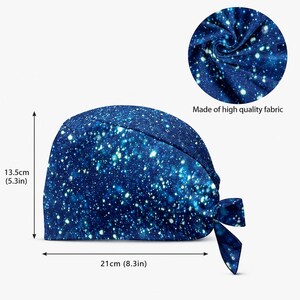 Op de afbeelding: Een blauwe operatiemuts met een witte sterren- en sterrenstelselprint. De muts is 13,5 cm hoog en 21 cm breed. Een cirkelvormige inzet toont de textuur van de stof met de tekst Made of high quality fabric onder de cirkel.