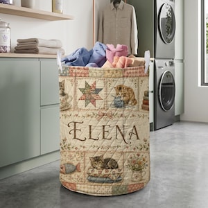 May include: A cylindrical quilted laundry hamper featuring a patchwork design with illustrations of cats, flowers, and stars. The name ELENA is printed in large brown script across the center. The hamper has two white fabric handles.