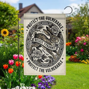 May include: A garden flag with the text "PROTECT THE VULNERABLE" and "RESIST TYRANNY" surrounding a detailed illustration of a rabbit. The flag is beige with black and white artwork, and is displayed in a garden setting.