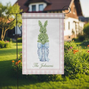 May include: A garden flag featuring a bunny topiary design. The bunny is made of green foliage and sits atop a blue and white vase, tied with a blue bow. The flag has a pink and white gingham border and the text "The Johnsons".