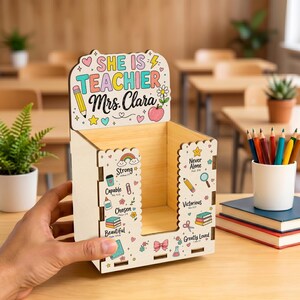 Könnte beinhalten: Ein Schreibtisch-Organizer aus hellem Holz mit der Aufschrift "SHE IS TEACHER Mrs. Clara" oben. Die Vorder- und Seitenflächen sind mit bunten Illustrationen von Stiften, Büchern, Herzen und Sternen sowie den Wörtern Strong, Capable, Chosen, Beautiful, Never Alone, Victorious und Greatly Loved verziert.