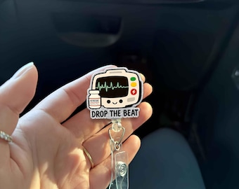Könnte beinhalten: Ein weißer und silberner Badgehalter mit einem Cartoon-Bild eines Herzmonitors mit einer Medizinampulle und dem Text "DROP THE BEAT".