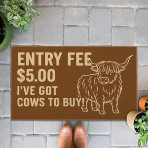 May include: Brown doormat with the text "ENTRY FEE $5.00 I'VE GOT COWS TO BUY!" and a drawing of a Highland cow. The mat is rectangular and sits on a gray tiled surface.