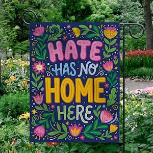 May include: A navy blue garden flag with the words "HATE HAS NO HOME HERE" in pink, yellow, and white. The flag is decorated with colorful floral and leaf designs. The flag is hanging in a garden.
