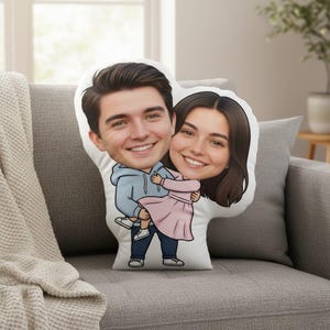 May include: A custom-shaped pillow featuring a cartoon illustration of a couple. The man is wearing a light blue hoodie and dark blue pants, holding the woman in a pink dress. The pillow is on a gray couch.