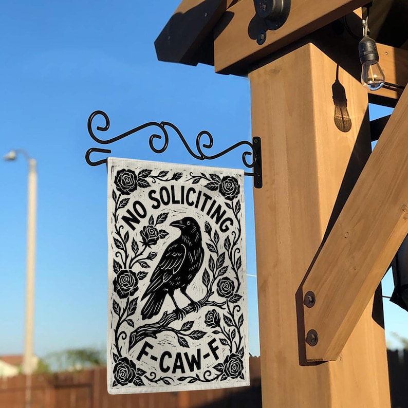 K&ouml;nnte beinhalten: Rechteckige Gartenflagge in Schwarz und Wei&szlig; mit einer Kr&auml;henillustration, Rosen und den Worten "NO SOLICITING" und "F-CAW-F". Die Flagge h&auml;ngt an einer schwarzen Metallhalterung an einem Holzpfosten.