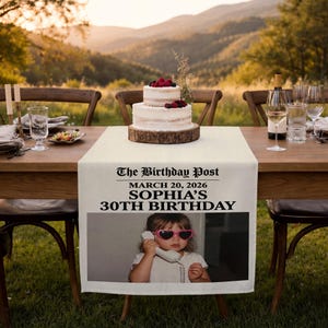 May include: A table setting with a two-tiered cake topped with berries on a wooden base. A cream-colored table runner displays the text "The Birthday Post" and "SOPHIA'S 30TH BIRTHDAY" with a photo of a child wearing heart-shaped sunglasses.