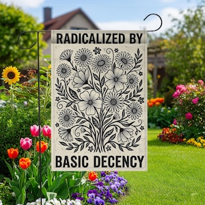 Op de afbeelding: Een tuinvaandel met de tekst "RADICALIZED BY BASIC DECENCY" boven en onder een zwart-wit bloemenontwerp. De vlag hangt aan een zwarte metalen standaard in een tuin met bloemen en groen.