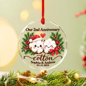May include: A clear circular acrylic ornament hanging from a red ribbon. It features a graphic of two smiling cotton bolls, one wearing a Santa hat, surrounded by holly and berries. Text reads: Our 2nd Anniversary, cotton, Sophia & Andrew, 03.23.2024.