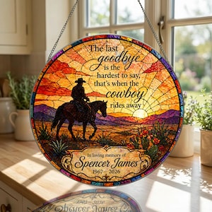 Puede incluir: Un adorno de ventana redondo de vidriera con un vaquero a caballo contra un atardecer. El texto dice: "El último adiós es el más difícil de decir, es cuando el vaquero se va." En memoria de Spencer James, 1967-2026.