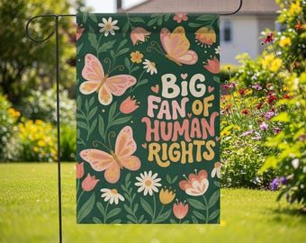 May include: A garden flag with a dark green background featuring butterflies, flowers, and the text "BIG FAN OF HUMAN RIGHTS" in pink, orange, and white. The flag is displayed outdoors in a garden.
