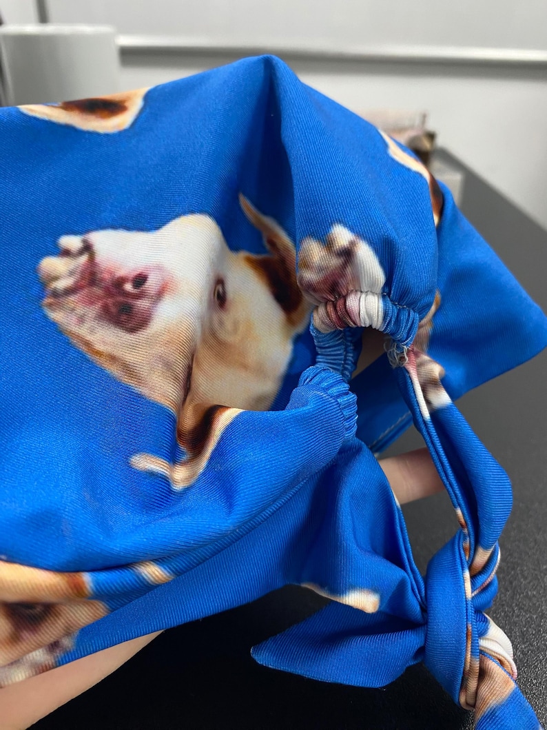 Op de afbeelding: Een felblauwe stoffen hoofddeksel met een herhalend patroon van een lichtbruine en witte hondenkop. Het accessoire is voorzien van een geplooid elastisch gedeelte en stoffen linten aan de achterkant voor een goede pasvorm.