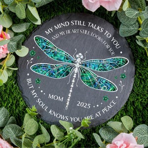 May include: A circular, dark grey stone featuring a dragonfly motif. The dragonfly has iridescent blue and green wings and a silver body. The text reads "MY MIND STILL TALKS TO YOU" and "BUT MY SOUL KNOWS YOU ARE AT PEACE". The word "MOM" and the year "2025" are also included.