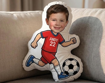 May include: A white, custom-shaped pillow featuring a cartoon illustration of a boy in a soccer uniform. The boy is wearing a red jersey with the name "ANDY" and the number "22". He is kicking a black and white soccer ball.