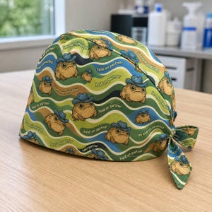 May include: A scrub cap featuring a repeating pattern of brown toads wearing blue cowboy hats against a background of wavy green and tan lines. The text "im overstimulated" and "hold on partner" is printed in small letters between the toads.