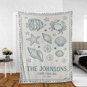 Puede incluir: Manta color crema con un diseño náutico azul que presenta un pez, un cangrejo, estrellas de mar, conchas marinas y dólares de arena. El texto "THE JOHNSONS, CAPE COD, MA, EST. 1994" está impreso en la parte inferior. La manta tiene un borde decorativo.