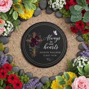 May include: A round black memorial stone with the text "Always in our hearts" and the name "Joseph Wagner" with dates. A gold cross with purple flowers is on the left. A butterfly is on the right. The stone is surrounded by flowers and rocks.