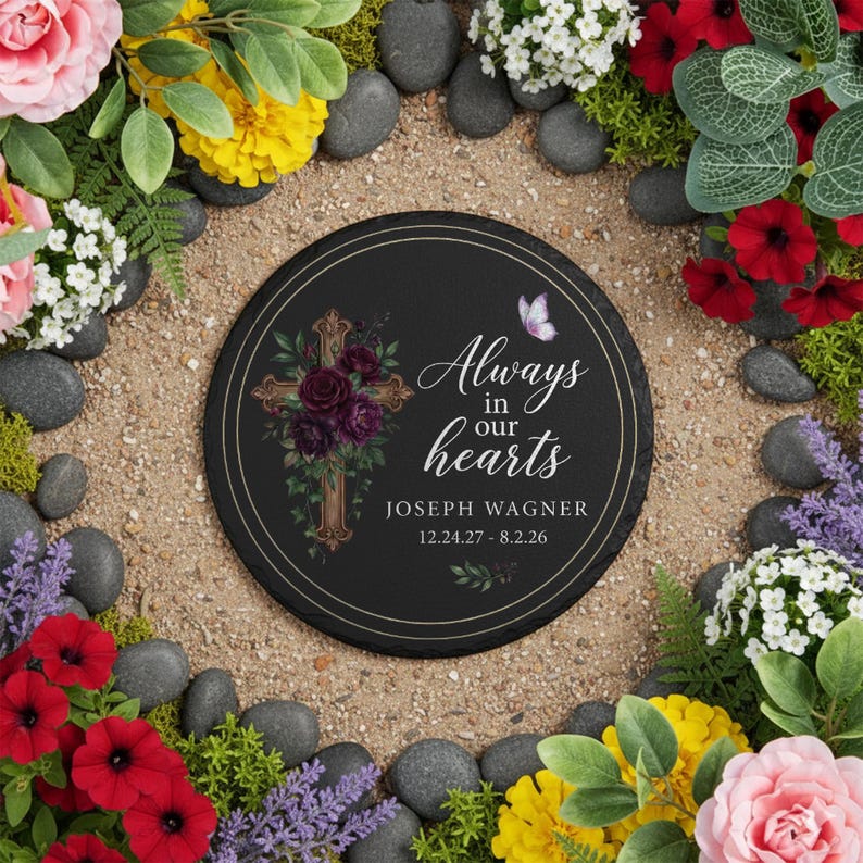 May include: A round black memorial stone with the text "Always in our hearts" and the name "Joseph Wagner" with dates. A gold cross with purple flowers is on the left. A butterfly is on the right. The stone is surrounded by flowers and rocks.