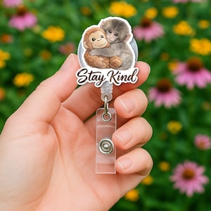 Pode incluir: Um porta-crachá com um design circular com dois macacos abraçados. O texto "Stay Kind" é exibido em uma fonte de script. O porta-crachá tem um corpo de plástico transparente com um fecho de metal e um cordão retrátil.