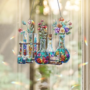 May include: A stained glass suncatcher featuring a collection of laboratory glassware including test tubes, beakers, and flasks filled with colorful flowers and greenery. A small periodic table is visible at the base of the glassware arrangement.