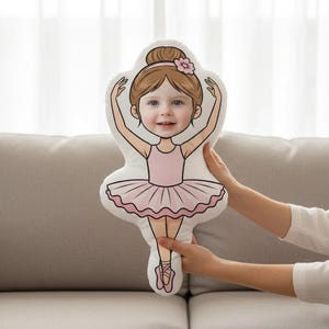 以下が含まれることがあります： バレリーナの形をしたカスタムデザインの枕。枕には、茶色の髪とピンクのチュチュを着た若い女の子の漫画イラストが描かれています。女の子の顔は写真です。枕は人が持っています。