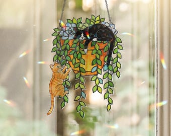 Puede incluir: Decoración colgante de ventana de vidrieras con un gato blanco y negro descansando en una maceta de color terracota con hojas verdes y flores azules. Un gato atigrado naranja trepa por las hojas. La decoración cuelga de una cadena.