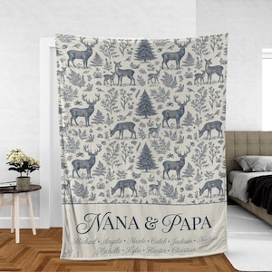 May include: A cream-colored blanket with a blue deer and forest pattern. The blanket has the words "NANA & PAPA" and several names printed on it. The blanket is displayed in a room with a bed and a side table.