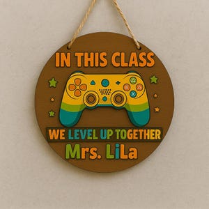 May include: A circular wooden sign with the text "IN THIS CLASS" and "WE LEVEL UP TOGETHER Mrs. LiLa". It features a yellow, green, and blue video game controller design, star accents, and a jute rope for hanging.